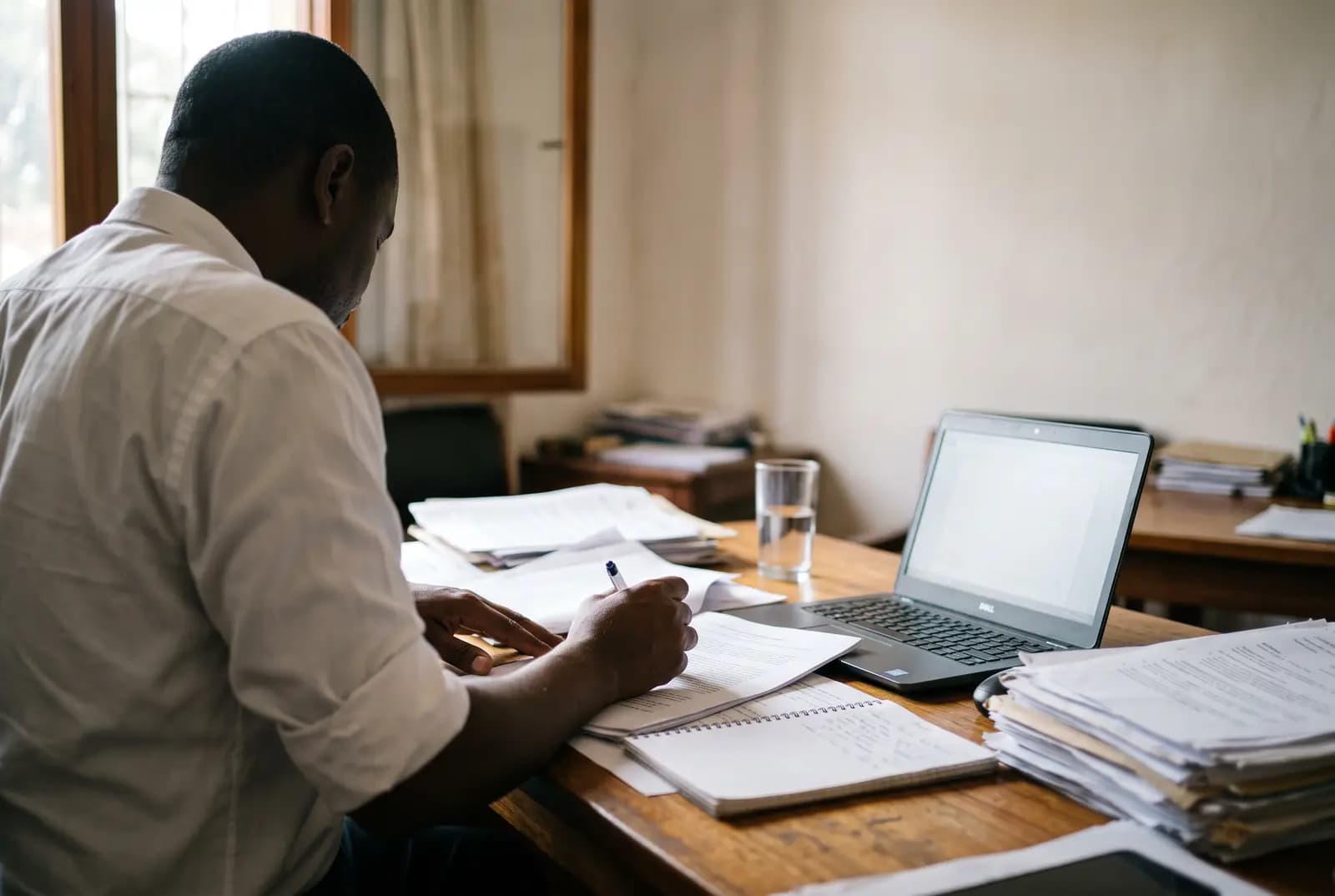 Un juriste penché sur un dossier à son bureau, stylo en main, dans la lumière d’une fenêtre ; papiers, carnet ouvert et ordinateur portable sur le plateau en bois.